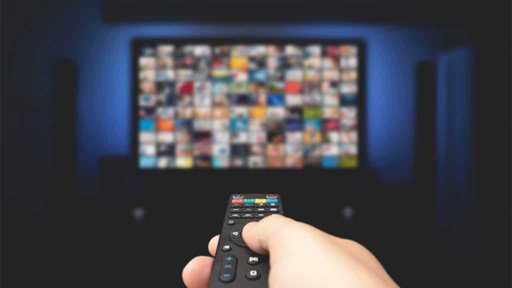 Pessoa apontando controle para TV lotada de imagens de programas; imagem ilustra conceito de TV 3.0