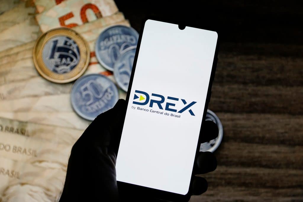 Imagem mostra, ao fundo, em uma mesa, notas de real e moedas; à frente, um smartphone com o logo do Drex na tela