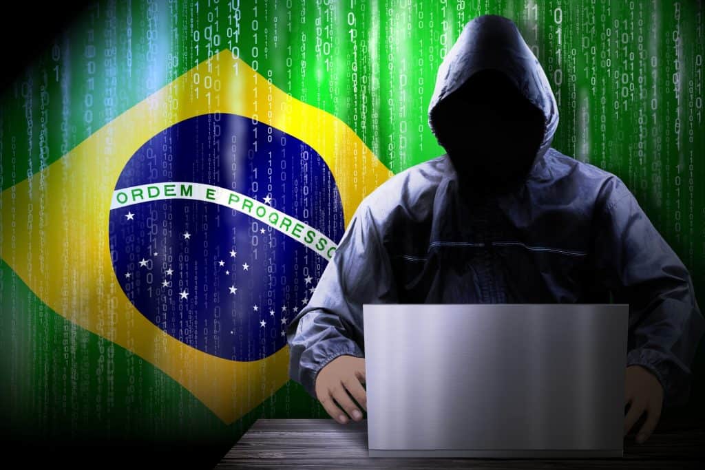 Pessoa na sombra com capuz atrás de um notebook e, ao lado, abbandeira do Brasil