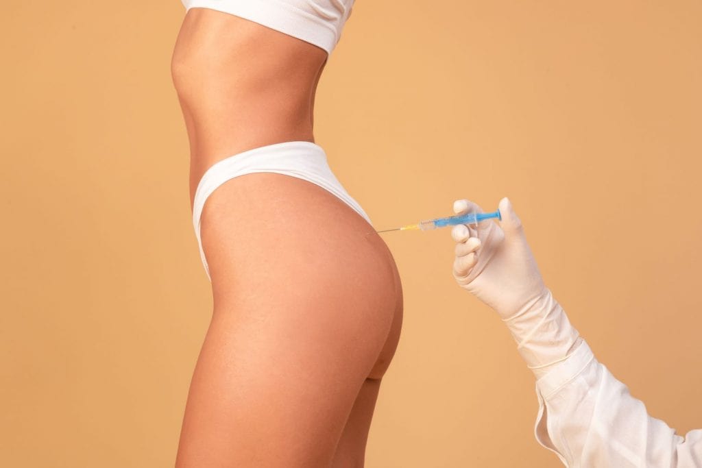 Conceito de lifting de bumbum não cirúrgico com Sculptra. Vista lateral de uma jovem recebendo injeção de quadril na região das nádegas, isolada em fundo de estúdio bege, recortada.