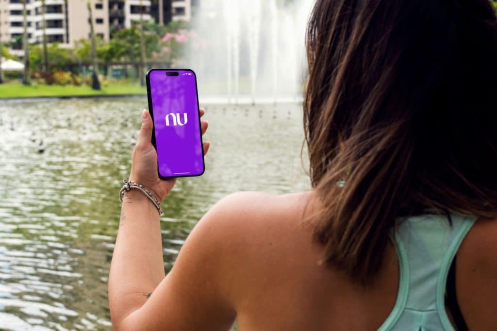Menina no parque segurando um smartphone iPhone 14 Pro com o app do Nubank na tela. Lago ao fundo. Rio de Janeiro, RJ, Brasil. Novembro de 2022.