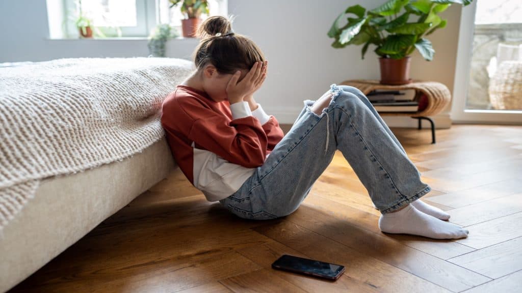 
Menina adolescente infeliz cobrindo o rosto com as mãos e chorando enquanto está sentada no chão com o celular perto, uma criança adolescente frustrada e chateada sendo assediada ou intimidada online. 

