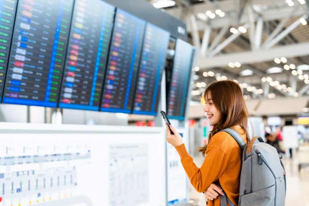
Jovem mulher asiática em aeroporto internacional, usando o smartphone e checando o voo no painel de informações.