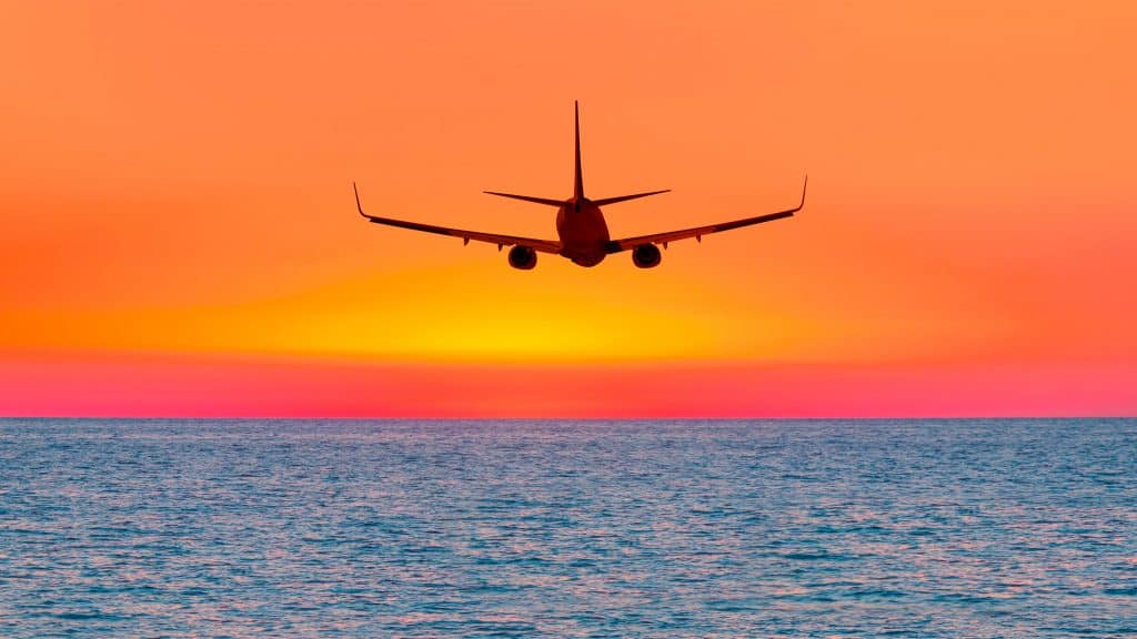 Silhueta da traseira de um avião sobrevoando o oceano durante com céu alaranjado ao fundo