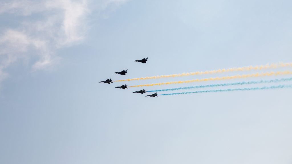 Seis caças F-16 voando em formação V soltando fumaça azul e amarela homenageando a Ucrânia