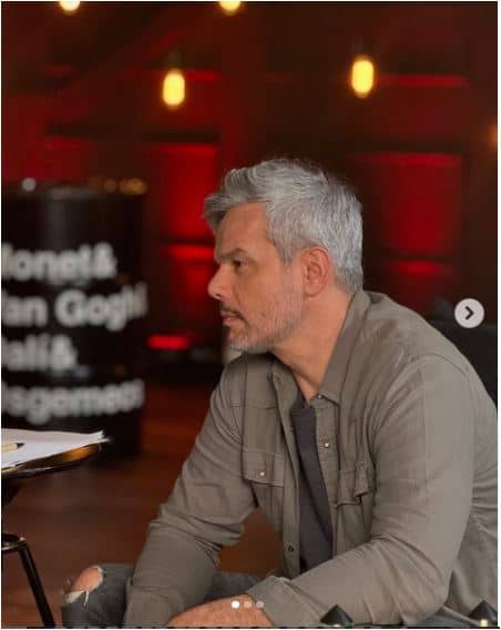 Homem bonito com cabelo grisalho, jaqueta marrom-claro e camiseta cinza, de perfil, em local com fundo vermelho.