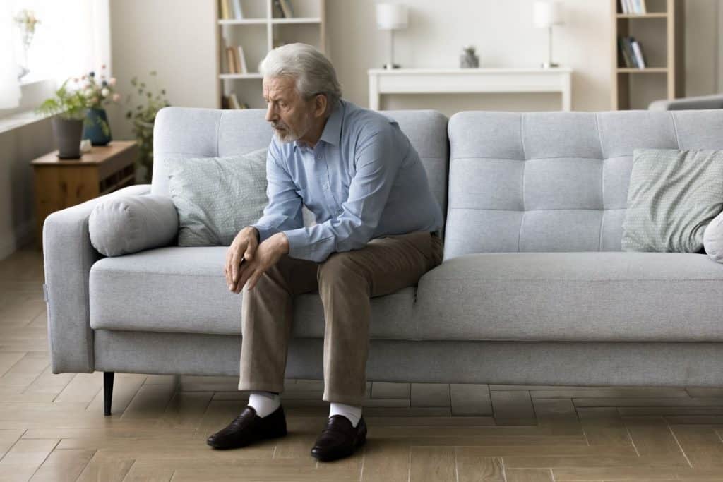Um homem idoso, solitário e triste, com cabelos grisalhos, está sentado sozinho em um sofá em casa. Ele olha para baixo, imerso em pensamentos negativos, sofrendo de depressão, apatia e lidando com perdas. Recebeu más notícias sobre saúde e problemas familiares