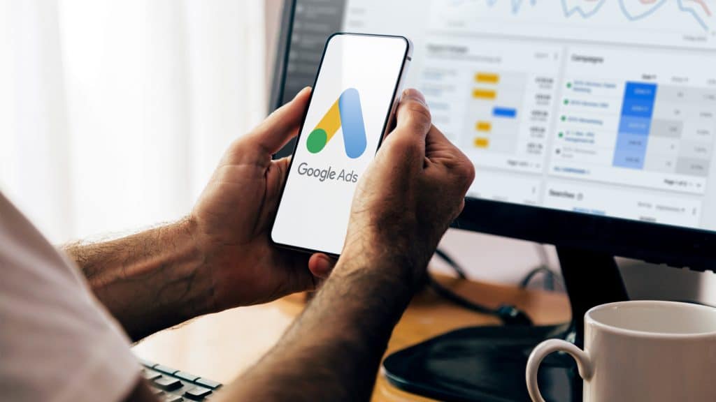 Como processo antitruste do Google pode mudar o mercado digital nos EUA O monopólio do Google no mercado de anúncios ainda está sob investigação. Homem segurando celular com logotipo do Google Ads na tela e, ao fundo , computador com gráficos no monitor