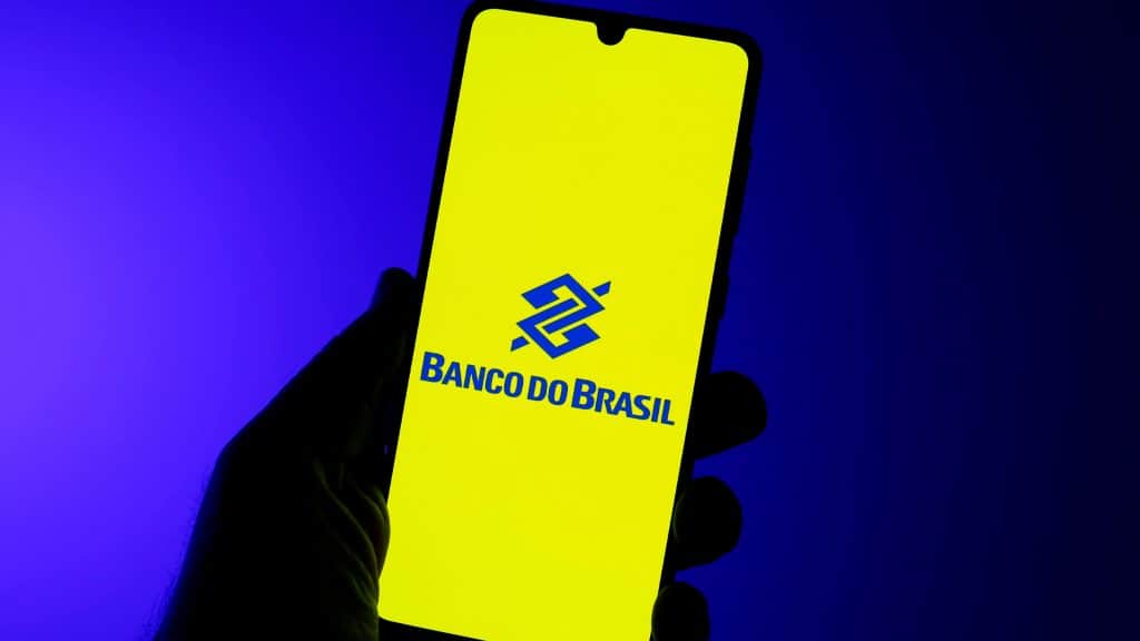 Pessoa segurando celular em frente fundo azulado com logomarca do Banco do Brasil na tela