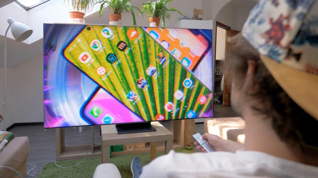Perspectiva sobre ombro de moço sentado assistindo vídeo sobre celulares em TV da Samsung
