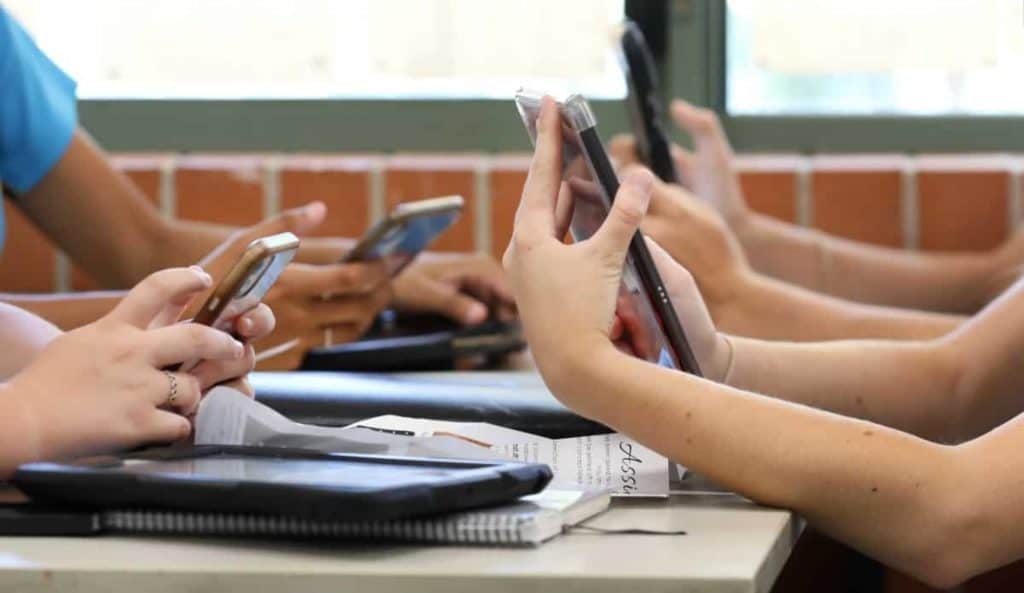 Mesa com alunos do ensino médio, todos segurando vários dispositivos digitais, como telefones e tablets, durante uma aula