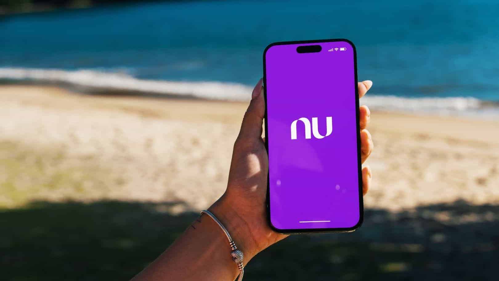 Garota na praia segurando um smartphone iPhone 14 Pro com o aplicativo do Nubank na tela. Rio de Janeiro, RJ, Brasil. 07 de junho de 2023.