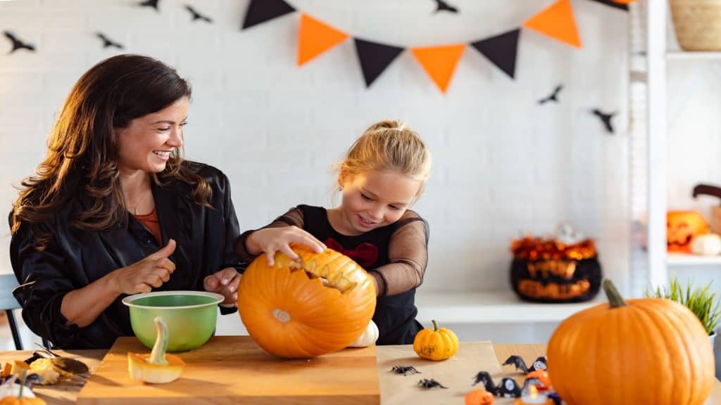 Uma mãe e sua filha sorriem enquanto esculpem abóboras juntas para o Halloween. A mesa está cheia de sementes de abóbora e decorações, e bandeiras festivas de Halloween penduradas na parede