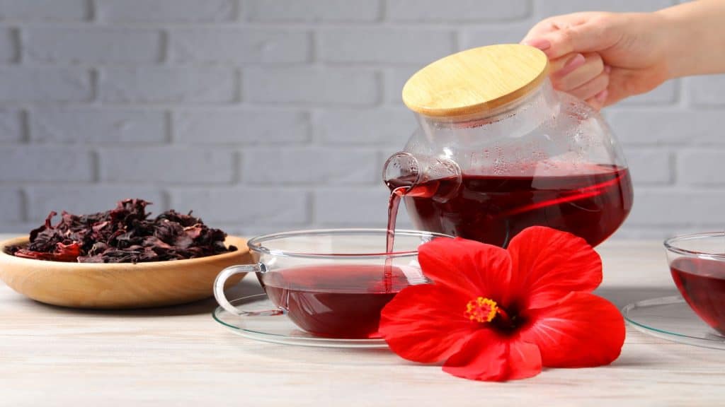 Mulher despejando delicioso chá de hibisco em uma xícara em uma mesa de madeira clara, close-up.