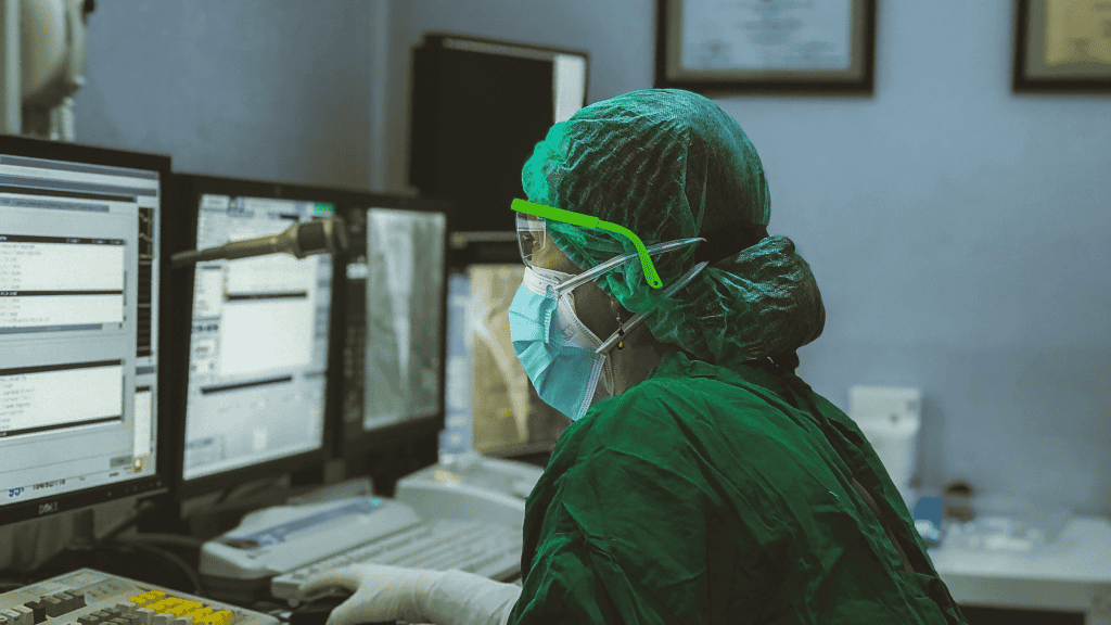 imagem mostra uma mulher paramentada com EPIs registrando um prontuário médico em um computador num hospital