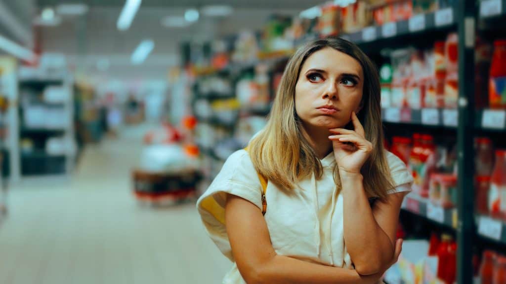 Cliente indeciso pensando o que comprar no supermercado. Mulher em dúvida pensa na inflação de preços e no consumismo.