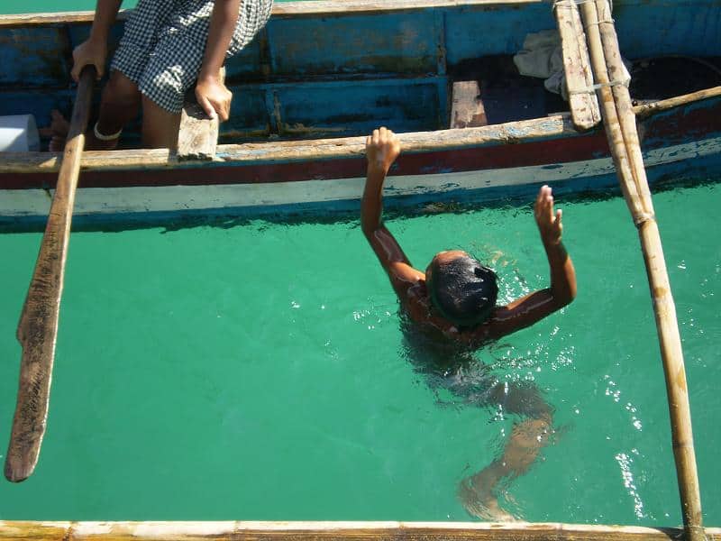 Povo Bajau: conheça os super-humanos que evoluíram para segurar o fôlego por 13 minutos dentro d’água Os seres humanos continuam a surpreender a ciência com sua capacidade de adaptação aos ambientes mais desafiadores do planeta. Apesar de muitos acreditarem que a evolução é algo que ocorreu apenas no passado distante, o exemplo do povo Bajau, também conhecido como “nômades do mar”, demonstra que a evolução continua a moldar o corpo humano até hoje. Uma criança Bajau mergulhando na água verde e cristalina, com os braços erguidos em direção à borda de um barco de madeira azul e branco, onde outra pessoa está sentada observando.