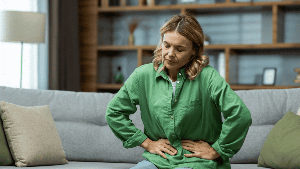Uma mulher loira está sentada no sofá em casa, segurando o estômago com as mãos. Sente a dor dos órgãos internos, indigestão, intoxicação, dor menstrual.
