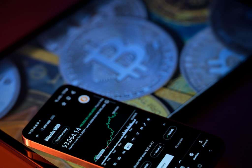 Vários logos de Bitcoin ao lado de um smartphone, que mostra gráficos