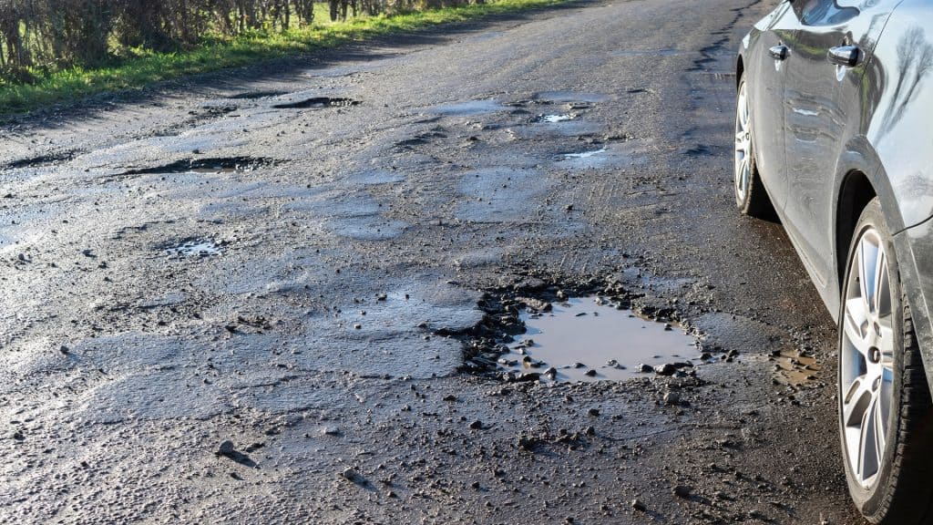 Um carro em uma estrada esburacada com buracos, asfalto destruído e poças. Poços de água em asfalto quebrado. Problemas de primavera e outono para os motoristas.