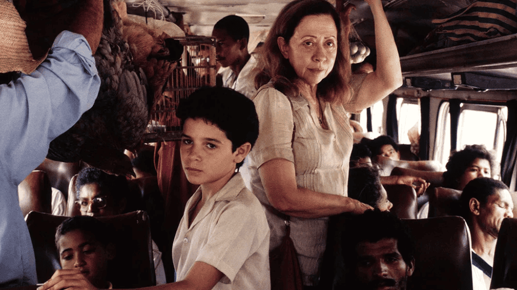 Uma mulher e um menino estão de pé em um ônibus lotado. Ela segura no suporte enquanto ele olha sério para a câmera. Passageiros e bagagens preenchem o espaço