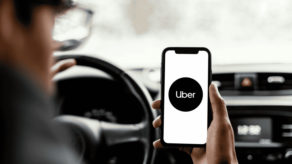 Motorista segura celular com o app da Uber aberto enquanto está ao volante, com interior do carro desfocado ao fundo