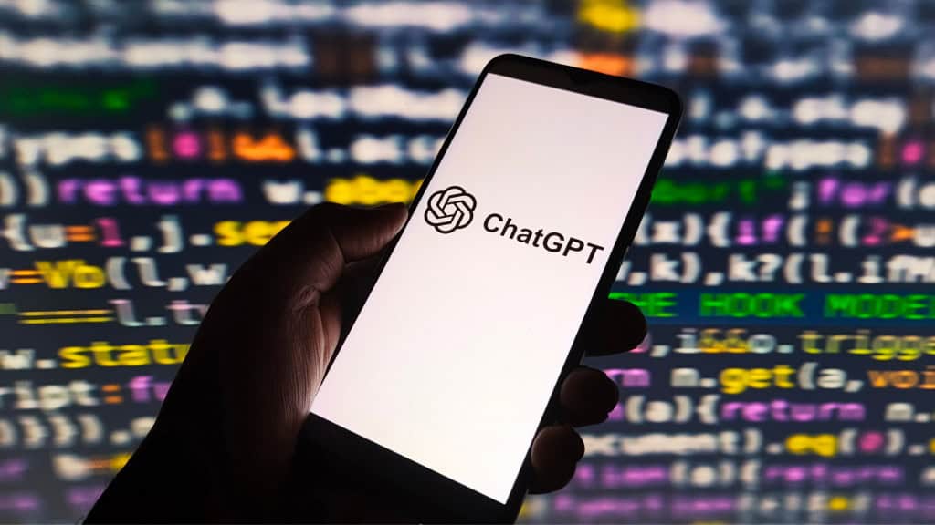 Pessoa segurando celular com logotipo do ChatGPT na tela; ao fundo, linhas de código de programação aparecem numa tela grande