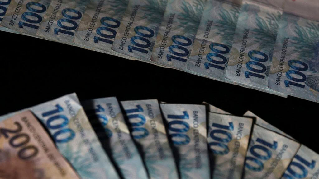 Notas de 100 e 200 reais organizadas em formatos de leque e fila numa mesa preta