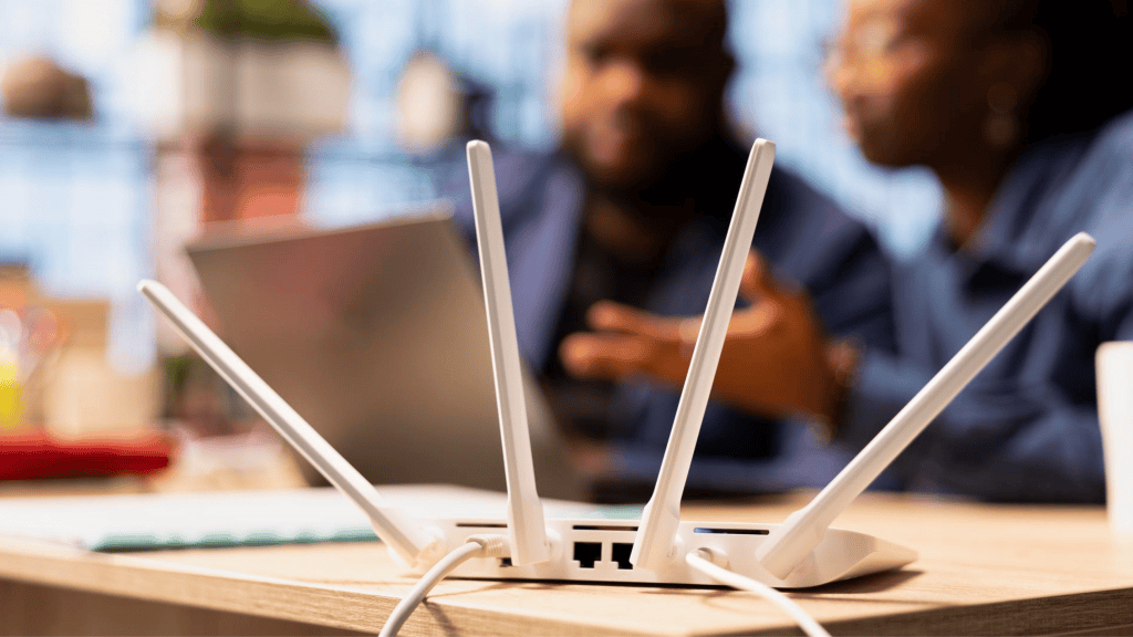 É seguro deixar modem ou roteador ligado direto? Por conta da praticidade de sempre ter internet quando precisar, muitas pessoas têm o costume de manter o modem ou roteador ligado o tempo inteiro, desligando-os ou reiniciando apenas em pequenos intervalos, como em momentos de falhas de sinal no aparelho. foto de um roteador em cima de uma mesa com pessoas e computadores atrás
