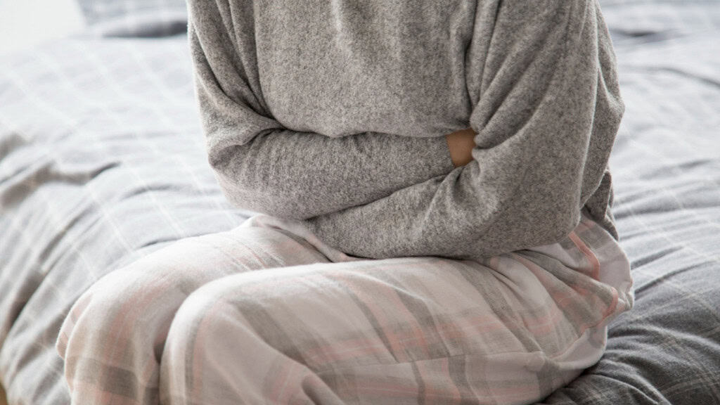 mulher de pijama sentada na cama enquanto aperta a barriga como se sentisse dor