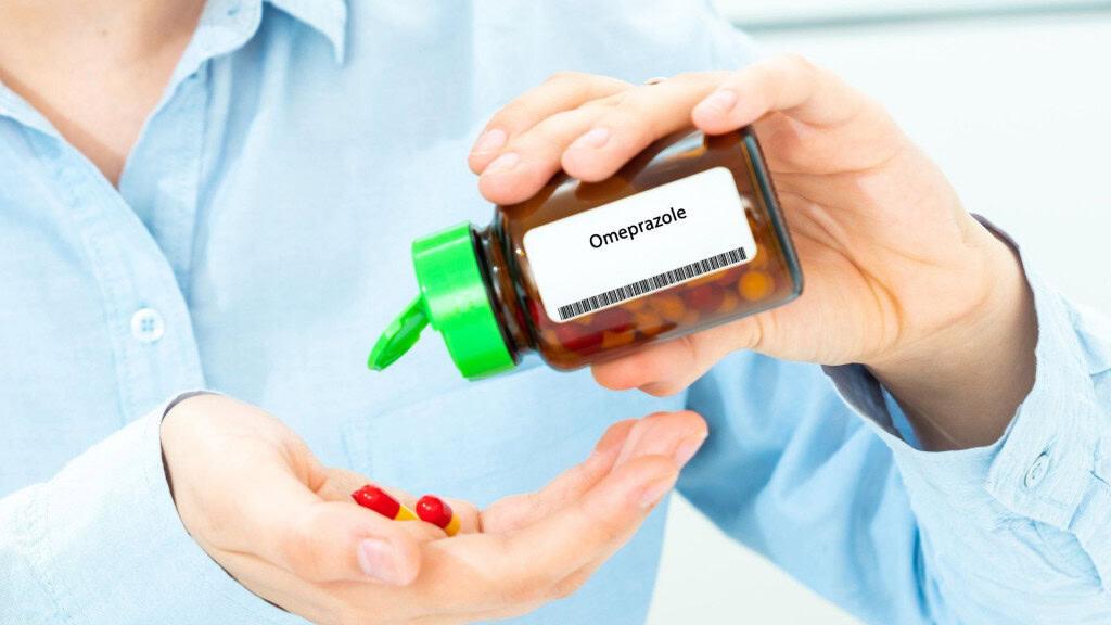 Pessoa com camisa azul segura um frasco marrom com tampa verde escrito “Omeprazol”.