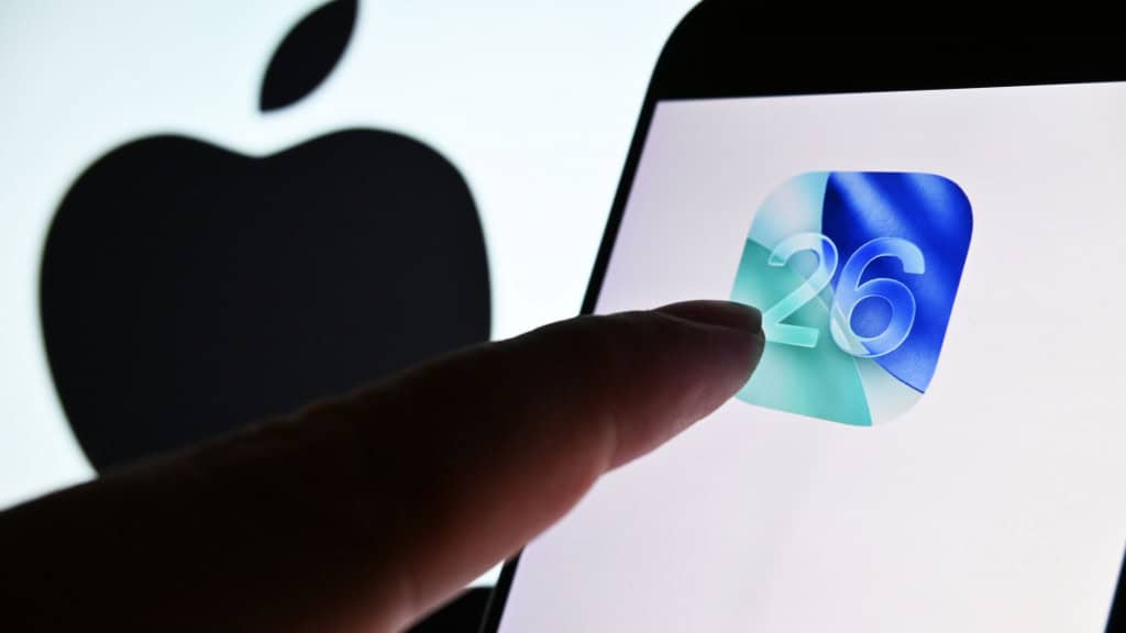 Apple prepara mudanças de segurança no iOS 26 O iOS 26 pode marcar um avanço importante na proteção de iPhones. Segundo informações do portal 9to5Mac, a Apple estaria preparando duas mudanças significativas em sua política de segurança, com foco em reduzir a exposição dos dispositivos a ameaças digitais. Dedo de pessoa quase tocando ícone do iOS 26 em tela de celular colocado na frente de tela maior exibindo o logotipo da Apple
