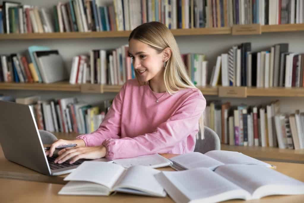 Menina estudando com livros abertos espalhados em uma mesa enquanto digita no notebook