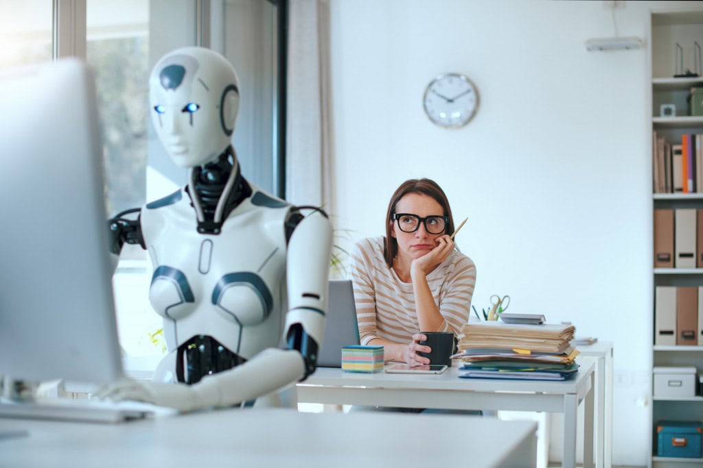 Do virtual para o real: como simulações podem revolucionar a inteligência artificial Os modelos de inteligência artificial atuais funcionam como alunos que passaram a vida inteira dentro de uma biblioteca virtual, mas nunca saíram para explorar a rua. Ou seja, esses modelos são bons em aprender com as informações já existentes – textos, imagens e vídeos –, mas não sabem lidar com situações do mundo real. Ao fundo, mulher com a cabeça apoiada na mão, com cara de tédio, olhando para um robô, que está à frente, trabalhando em um computador