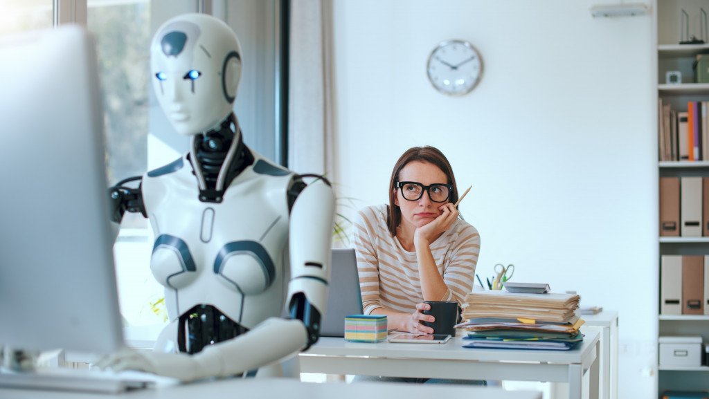CEO do Zoom acredita que IA pode reduzir semana de trabalho Apesar dos temores relacionados aos cortes de empregos, a inteligência artificial tem acelerado alguns processos e ajudado a mudar o mercado de trabalho como conhecemos hoje. No entanto, mais mudanças podem acontecer no futuro. Ao fundo, mulher com a cabeça apoiada na mão, com cara de tédio, olhando para um robô, que está à frente, trabalhando em um computador