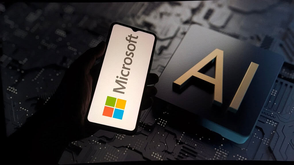 Pessoa segurando celular com logotipo da Microsoft na tela na frente de outra tela, esta grande, exibindo ilustração de chip de inteligência artificial (IA)
