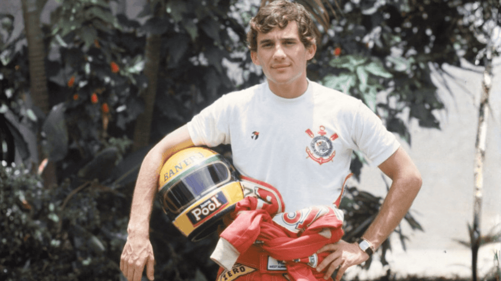 Ayrton Senna de camiseta branca do Corinthians segura um capacete de corrida amarelo com faixas verde e azul, posando ao ar livre ao lado de plantas.