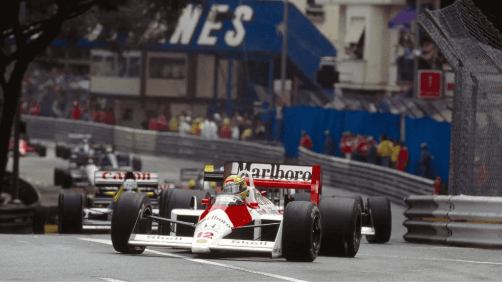 Por que Ayrton Senna é considerado o rei do GP de Mônaco na Fórmula 1? Nenhum outro piloto deixou tamanha marca em Monte Carlo quanto Ayrton Senna. Em uma pista onde cada milímetro conta, o brasileiro alcançou um feito inédito e ainda insuperável: seis vitórias no GP de Mônaco, sendo cinco delas consecutivas. Seu domínio no traçado urbano não se resumiu a números, mas a uma conexão singular entre piloto, máquina e paixão. Carro de Fórmula 1 da McLaren, pilotado por Ayrton Senna, lidera corrida nas ruas estreitas do GP de Mônaco, seguido por outros carros.