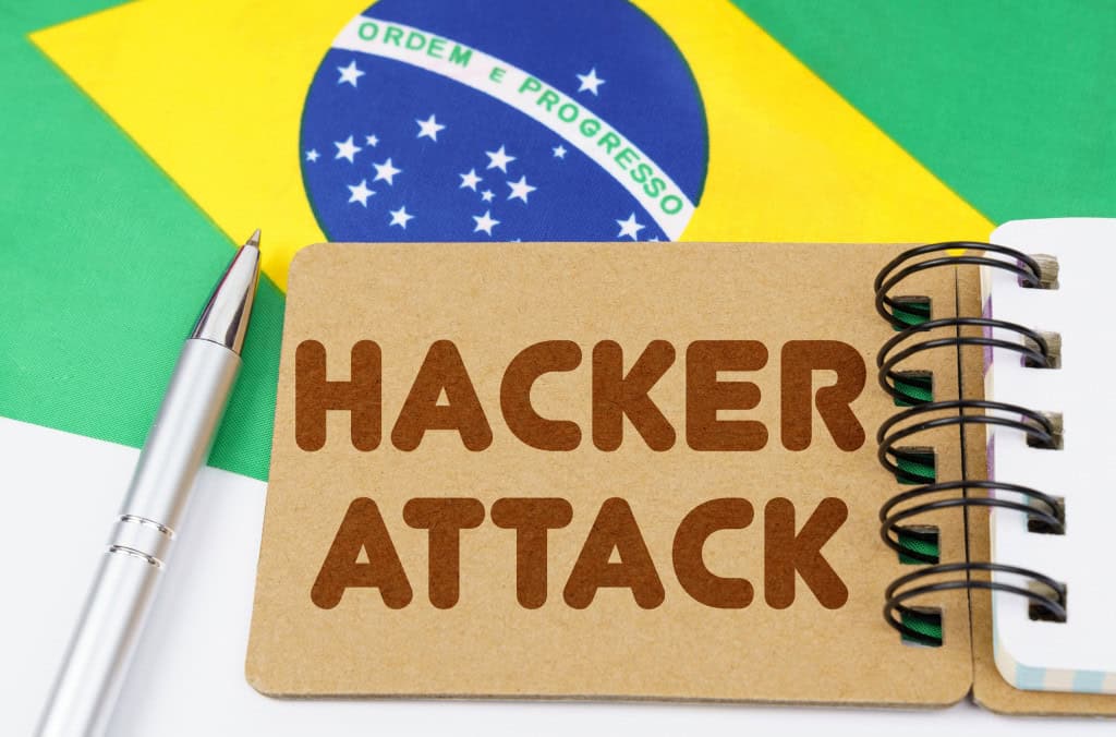 PF vai às ruas em operação contra grupos de ataques cibernéticos Nesta sexta-feira (17), a Polícia Federal (PF) foi às ruas para deflagrar a Operação Decrypt, que possui dois mandados de busca e apreensão em Minas Gerais, outro de busca e apreensão e um de prisão temporária em São Paulo. O intuito da operação é investigar a suposta participação de um brasileiro em uma organização criminosa transacional que realiza ataques cibernéticos via ransomware. Ao fundo, uma bandeira do Brasil em cima de uma mesa branca; em cima dela, uma caneta e um bloco de notas; No bloco de notas, lê-se "hacker attack"