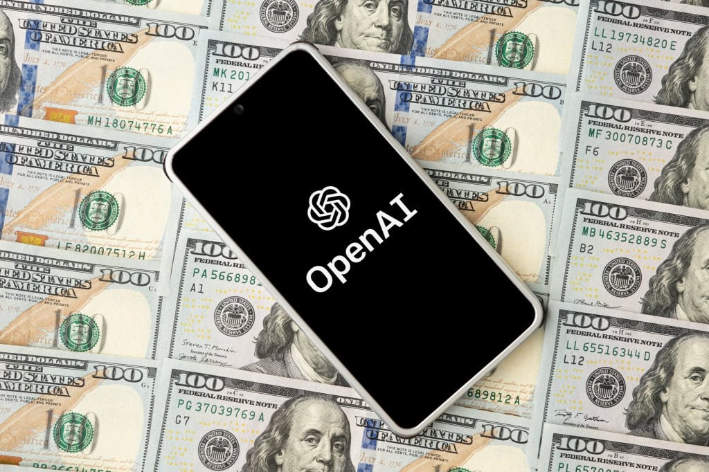 OpenAI: como Sam Altman está “reinventando” finanças para bancar IA A ascensão bilionária da OpenAI, liderada por Sam Altman, tem sido impulsionada não apenas por inovações tecnológicas, mas, também, por um complexo e criativo sistema de financiamentos. Logo da OpenAI em smartphone que está em cima de notas de dólar