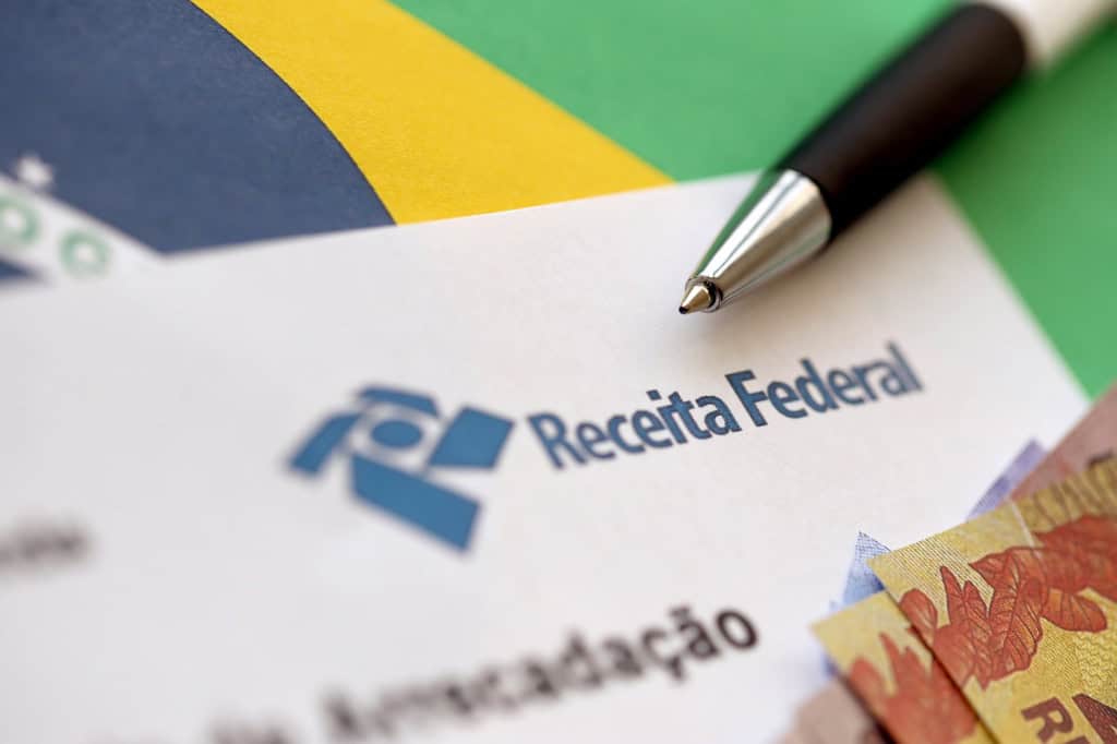 Ao fundo, bandeira do Brasil; em cima, um papel com o logo da receita federal, uma caneta e algumas notas de real