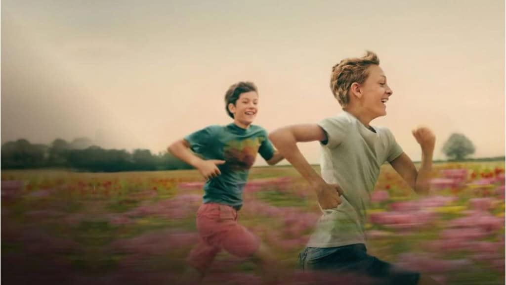Dois adolescentes correm sorridentes em meio a uma plantação com flores rosas ao fundo.