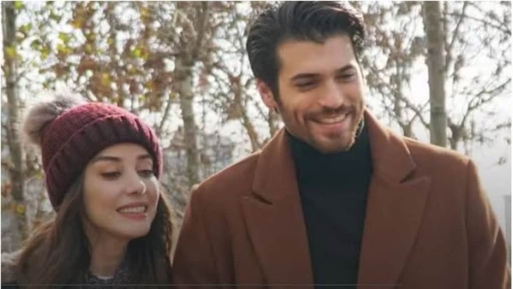 Duas pessoas sorridentes estão em meio a árvores sem folhas; uma usa gorro vinho e casaco escuro, a outra veste sobretudo marrom e gola alta preta.