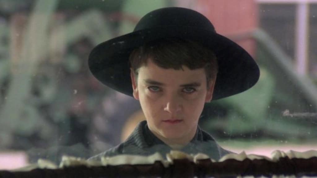 menino de olhos claros e expressão malévola, usando chapéu