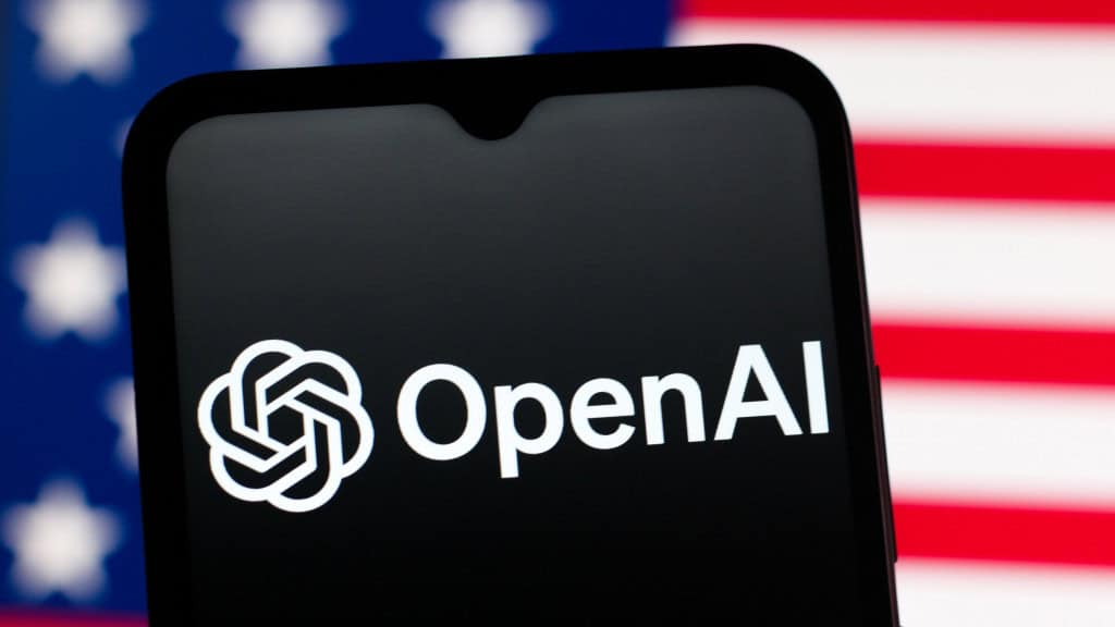 Celular com logotipo da OpenAI na tela colocado na frente de tela exibindo bandeira dos Estados Unidos