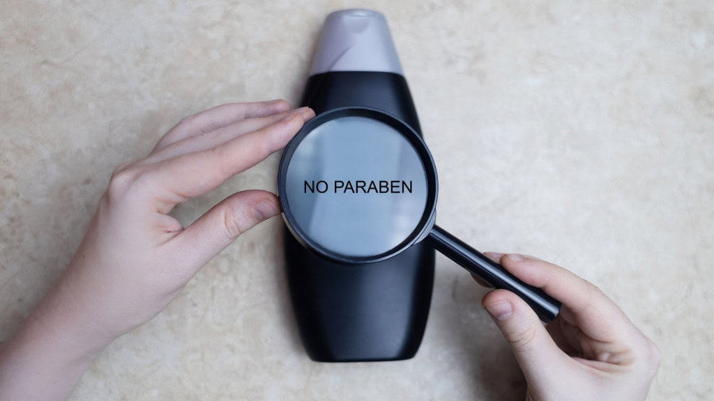 shampoo ou sabonete líquido, apoiado sobre uma superfície clara. Duas mãos seguram uma lupa posicionada sobre o frasco. A lupa amplia uma parte do rótulo, revelando em letras brancas a frase “NO PARABEN”.
