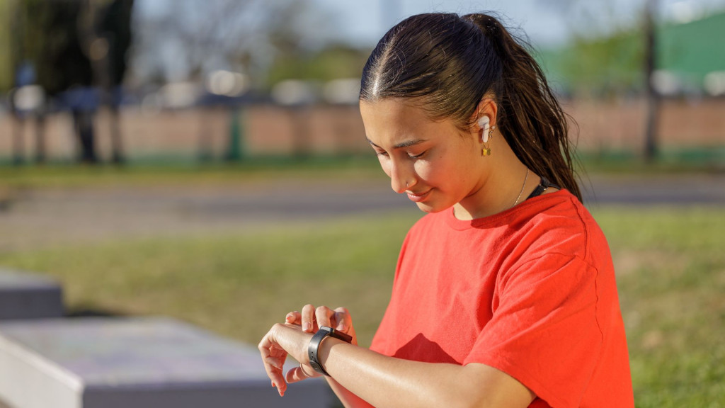 Mulher com camisa vermelha olhando seu smartwatch sentada em um banco, usando fones sem fio