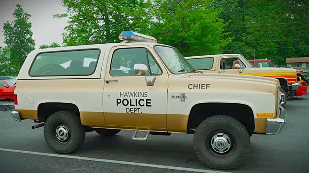 Imagem de uma cena de Stranger Things mostrando um Chevrolet K5 Blazer 1983 laranja ou marrom clássico em uma estrada em Hawkins
