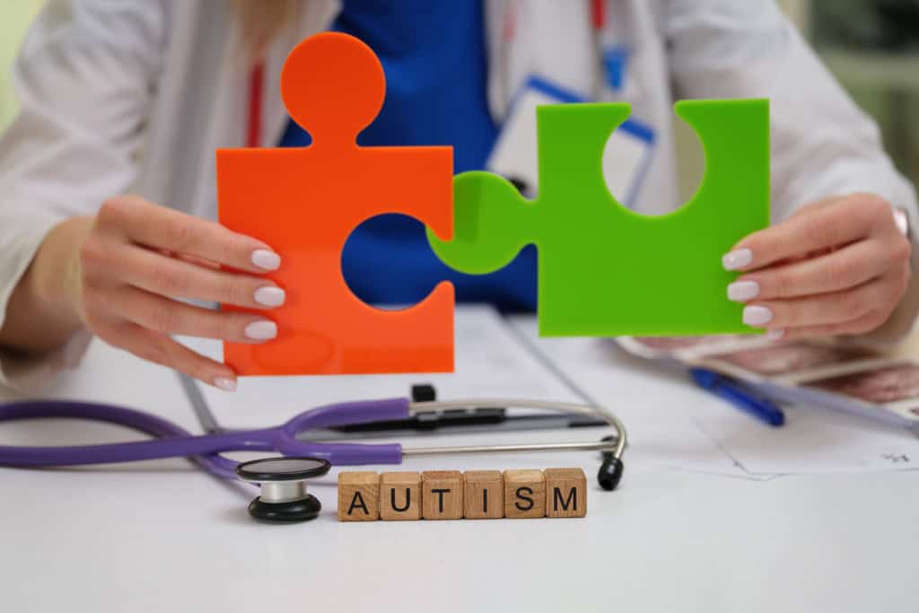 Imagem mostra mãos de médica sentada à mesa segurando peças coloridas de quebra cabeça com a palavra autism visível, representando a avaliação clínica usada para identificar sinais de autismo