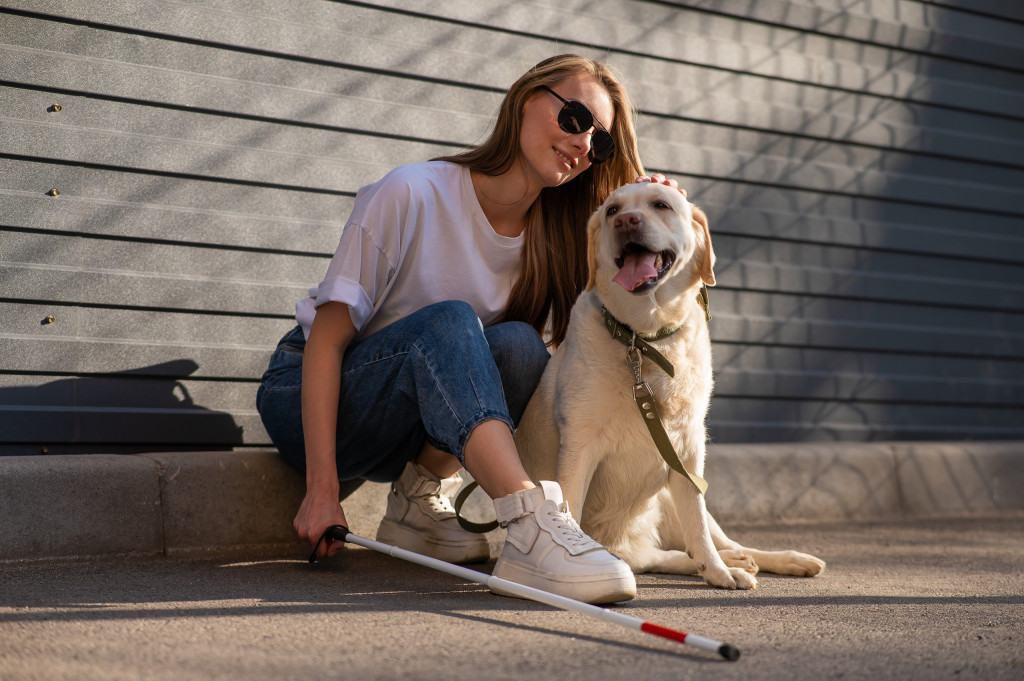 Uber disponibiliza ferramenta para deficientes visuais e cães-guia A Uber anunciou um novo recurso que promete ajudar bastante os usuários com deficiência visual e que possuem cães-guia. A ideia é evitar constrangimentos ou recusas de motoristas, dando autonomia ao usuário. Famílias socializadoras (que “treinam” o cão-guia) também poderão utilizá-lo. Mulher loira deficiente visual sentada no meio-fio, de óculos e segurando uma bengala na mão direita; à sua esquerda, está um cão-guia; ela está acariciando o cão